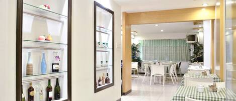 A hotel corridor with a glass display case on the left, a dining area with checkered tablecloths on the right, and a television mounted on the wall.