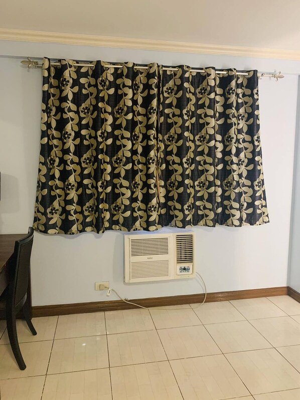 A room with a window covered by a black and gold patterned curtain, a wall-mounted air conditioner, and a tiled floor. - Jupiter Suites (Makati)