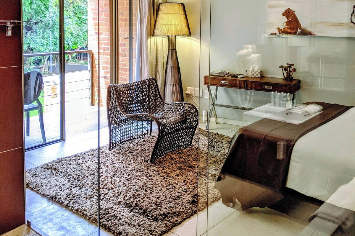 A modern hotel room with a glass door leading to a balcony, a bed with a white headboard, a small desk with a lamp, and a television mounted on the wall.