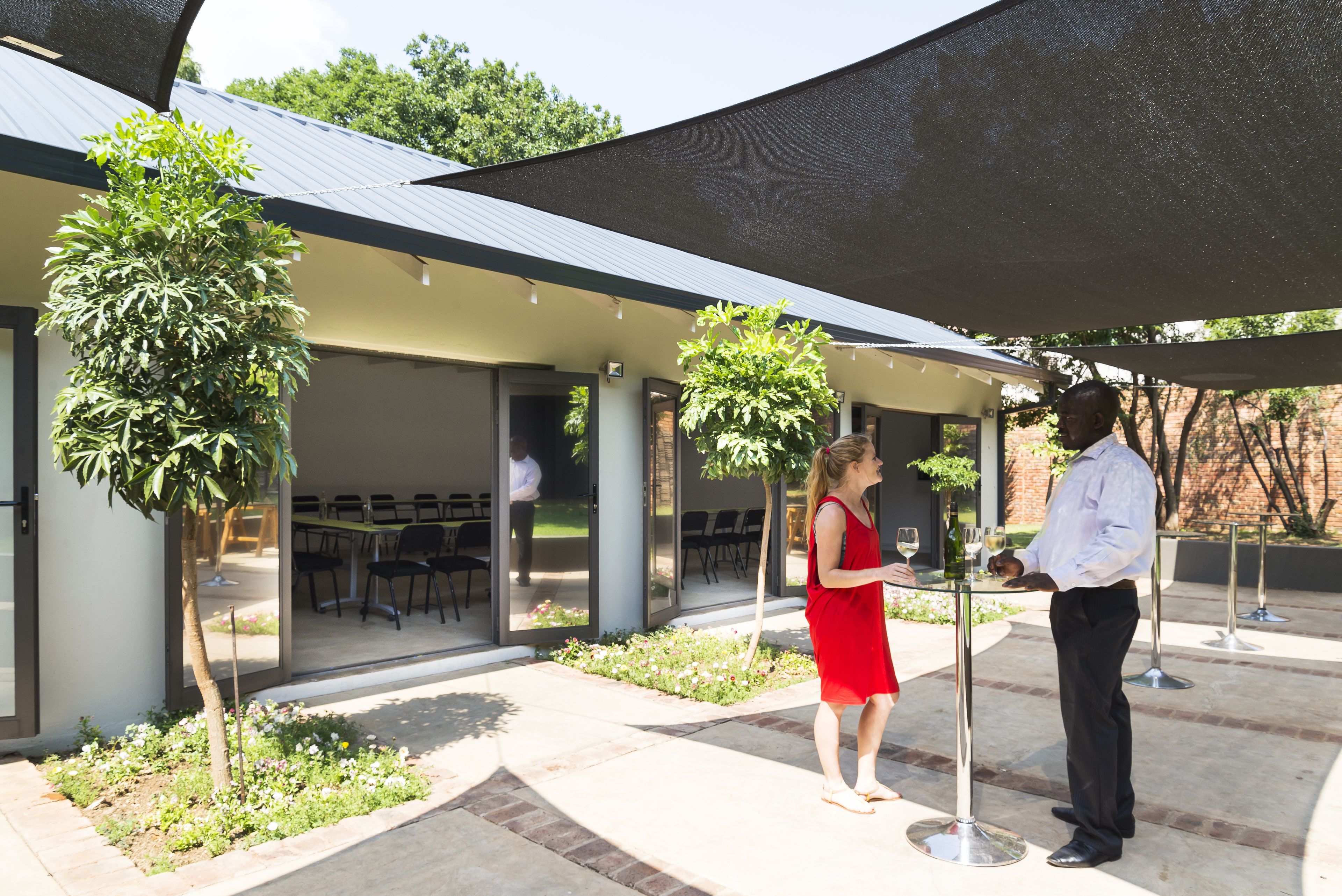 An outdoor patio area with a covered seating area, a small garden, and a waiter serving a guest.