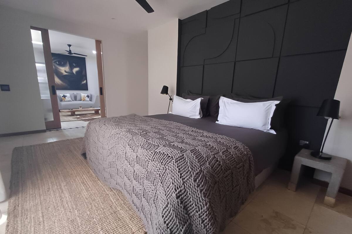 A modern bedroom with a large bed, a dark headboard, a bedside table with a lamp, and a view of a living area with a sofa and a painting.