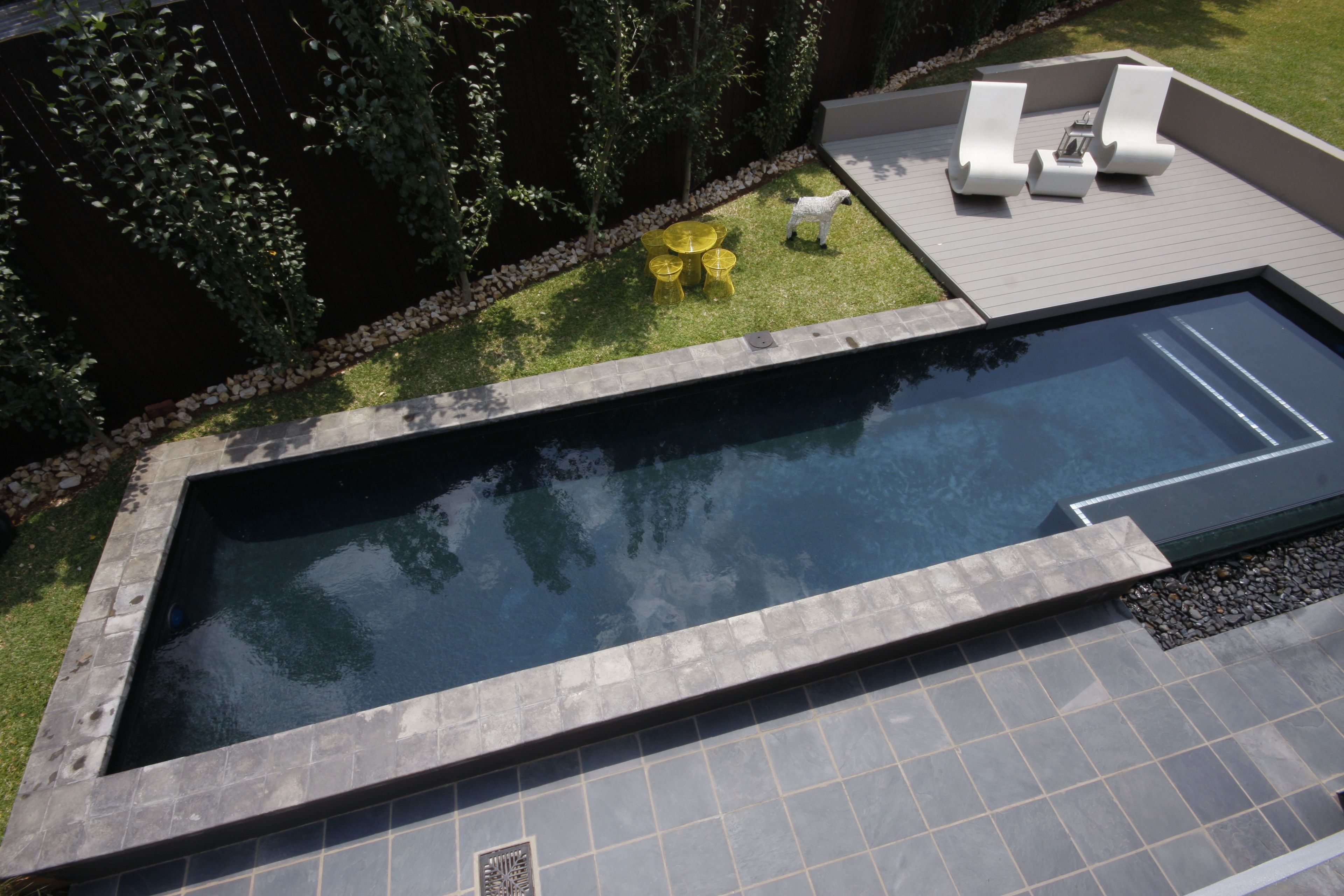 A rectangular swimming pool with a tiled deck, surrounded by a grassy area, a small seating area with white lounge chairs, and a tree.