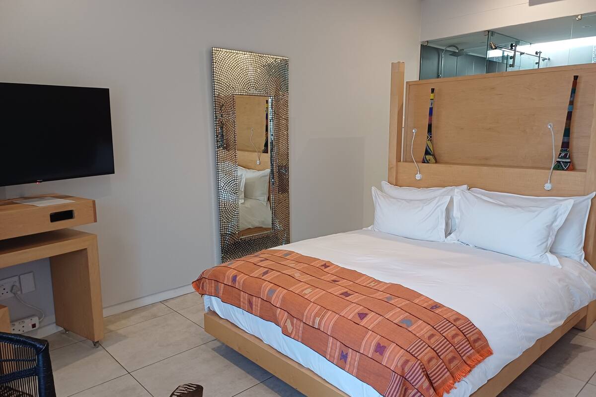A hotel room with a bed, a TV, a wooden headboard, a large mirror, and a small wooden table.