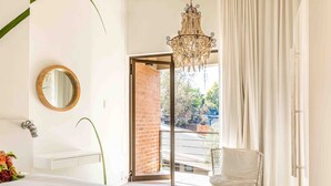 A bedroom with a bed, a round mirror, a chandelier, a white sink, and a view of a brick wall and greenery outside. - Alpine Attitude Boutique Hotel & Conference Venue (Pretoria)