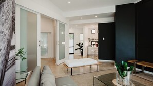 A modern living room with a sofa, a glass coffee table, and a bench. There is a kitchen area in the background with a dining table and chairs. - Alpine Attitude Boutique Hotel & Conference Venue (Pretoria)