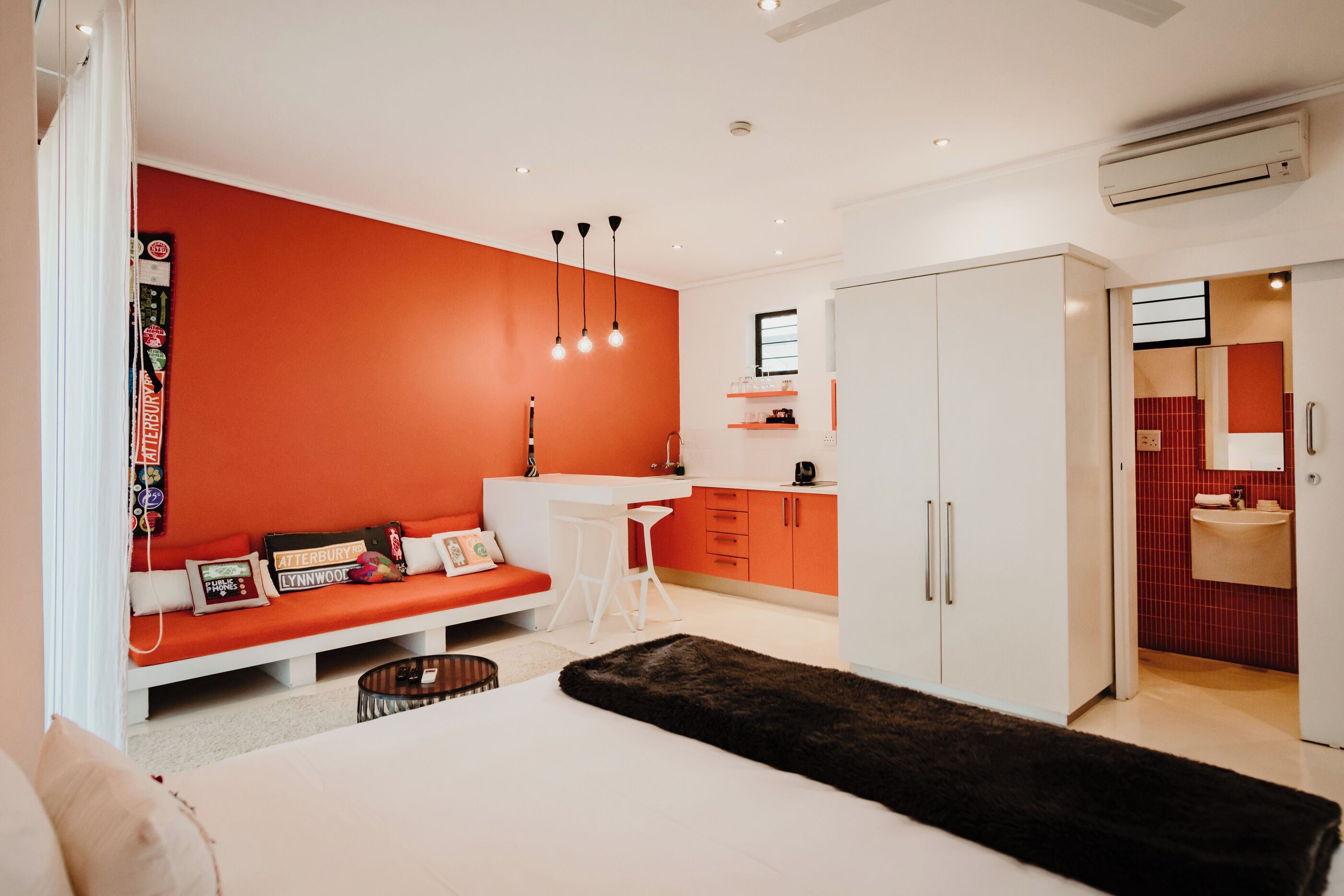 Une chambre à coucher moderne avec un grand lit, une armoire blanche, un petit bureau et un mur d’accent rouge.