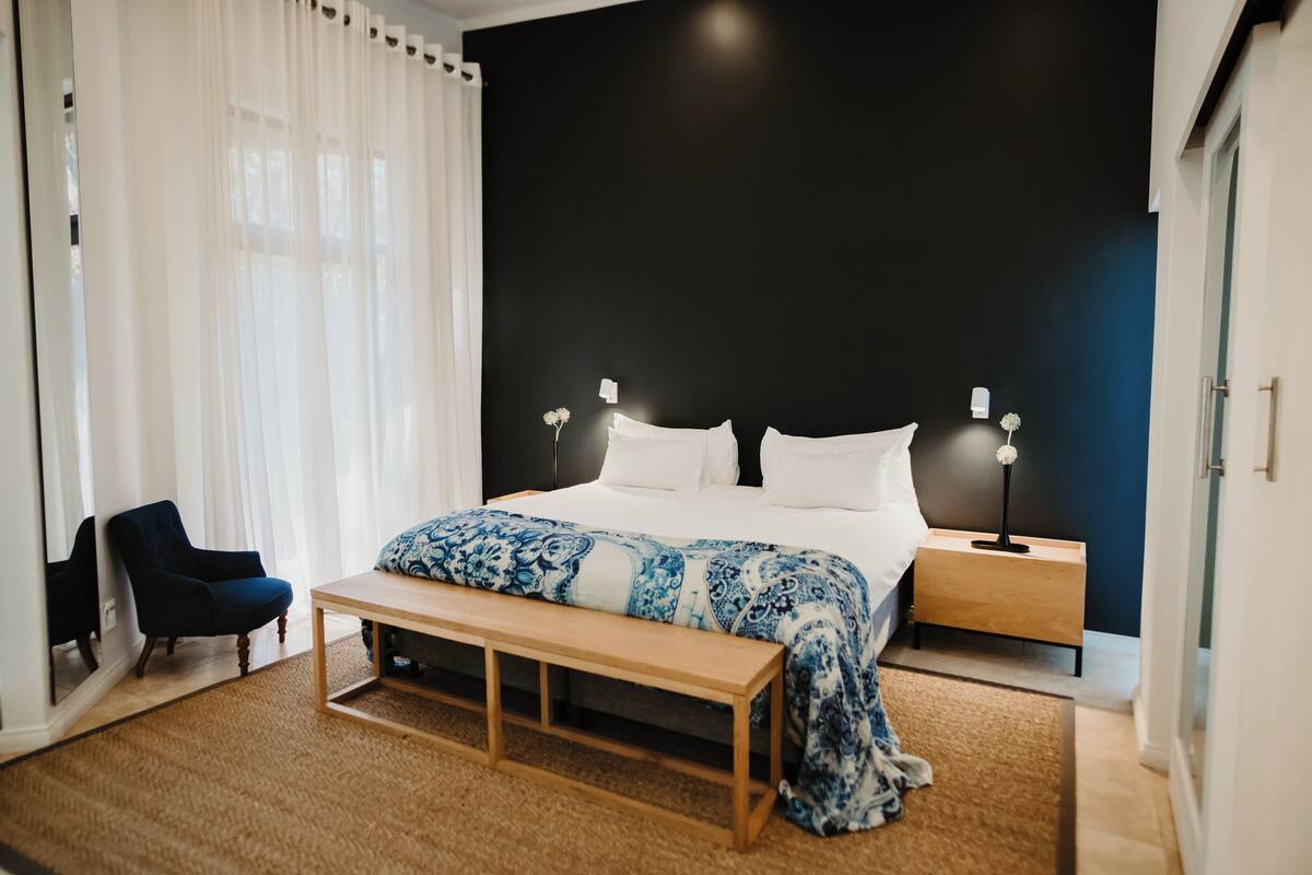 A bedroom with a large bed, a bench, a chair, and a nightstand with a lamp.