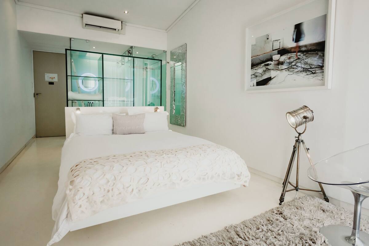 A modern bedroom with a large bed, a glass shower enclosure, a mounted picture, and a tripod lamp.