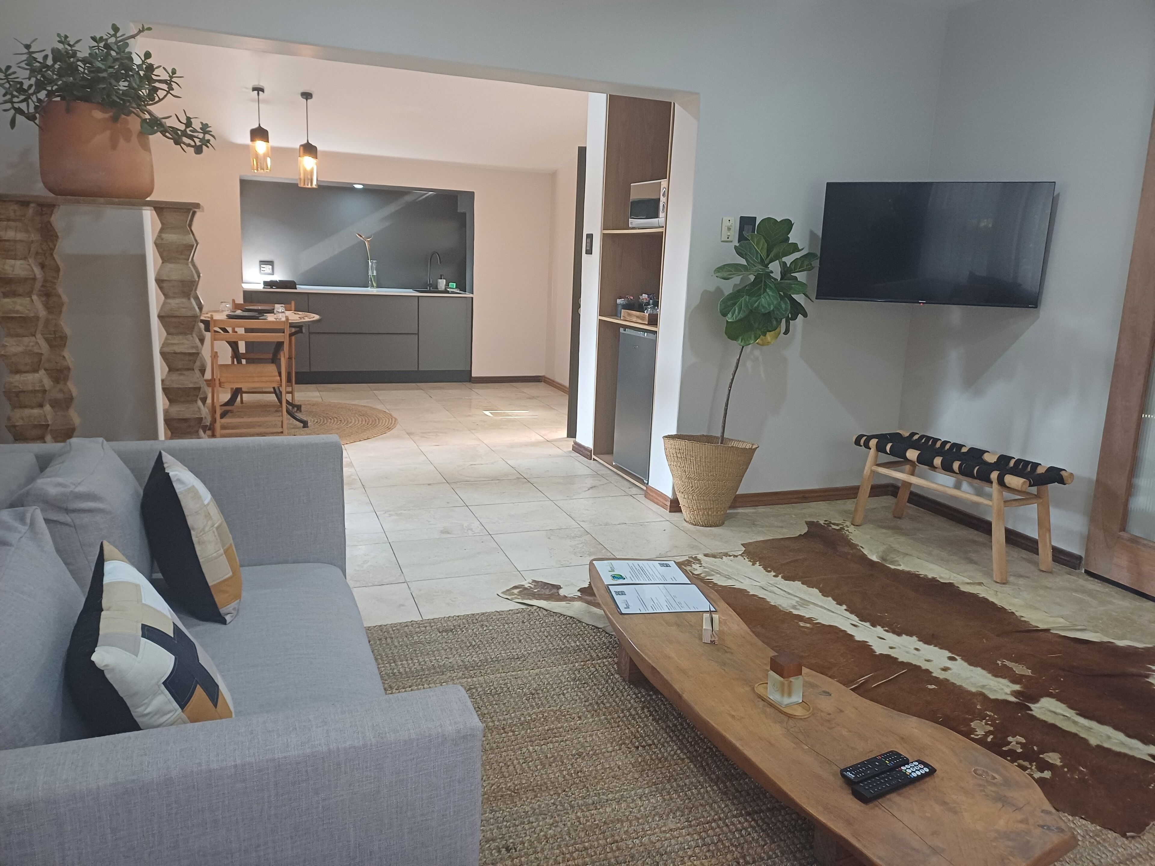 A living room with a sofa, a coffee table, a TV mounted on the wall, a wooden bench, and a kitchen area in the background.