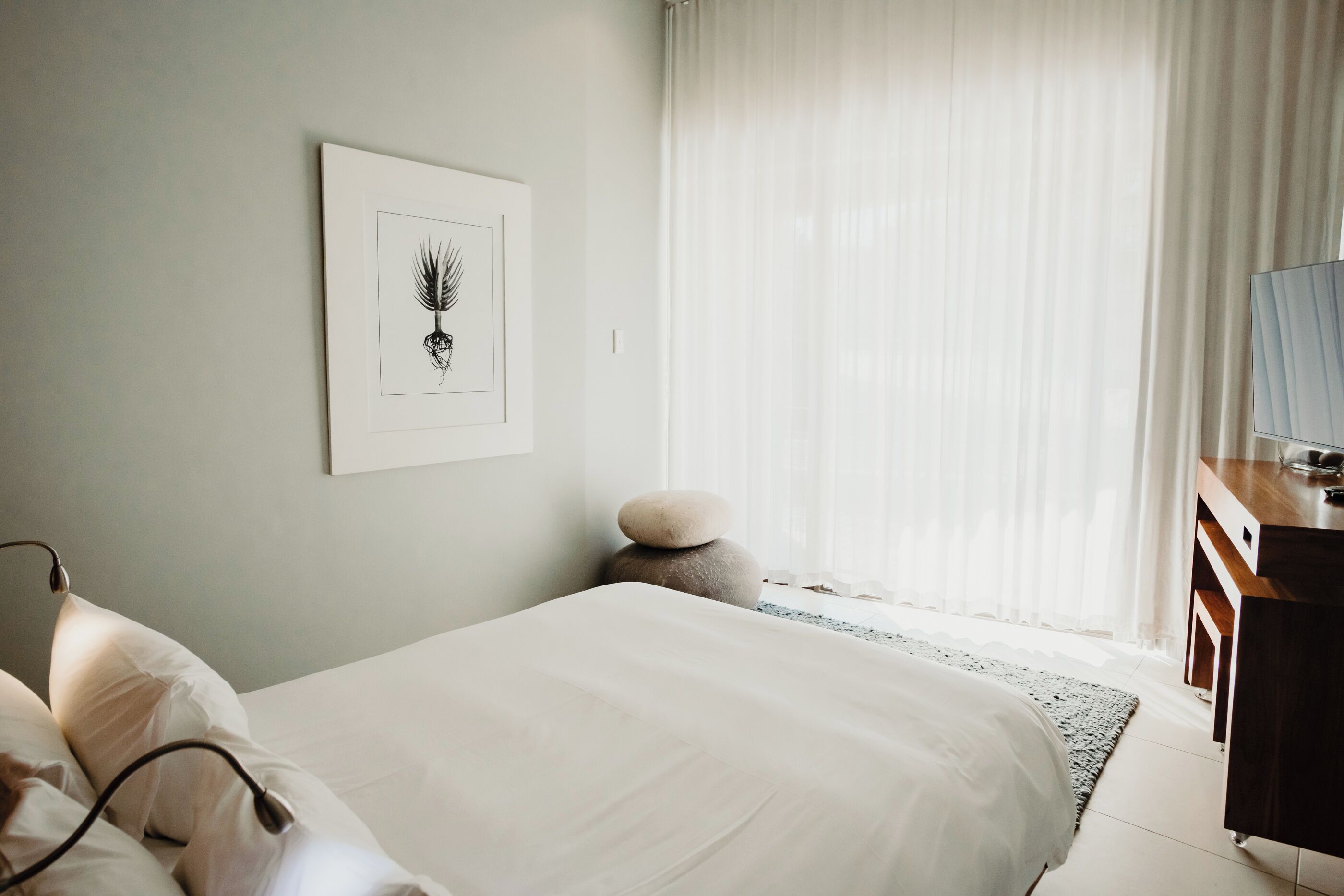Un dormitorio con una cama, una mesita de noche, una lámpara, una obra de arte enmarcada en la pared y una ventana con cortinas.