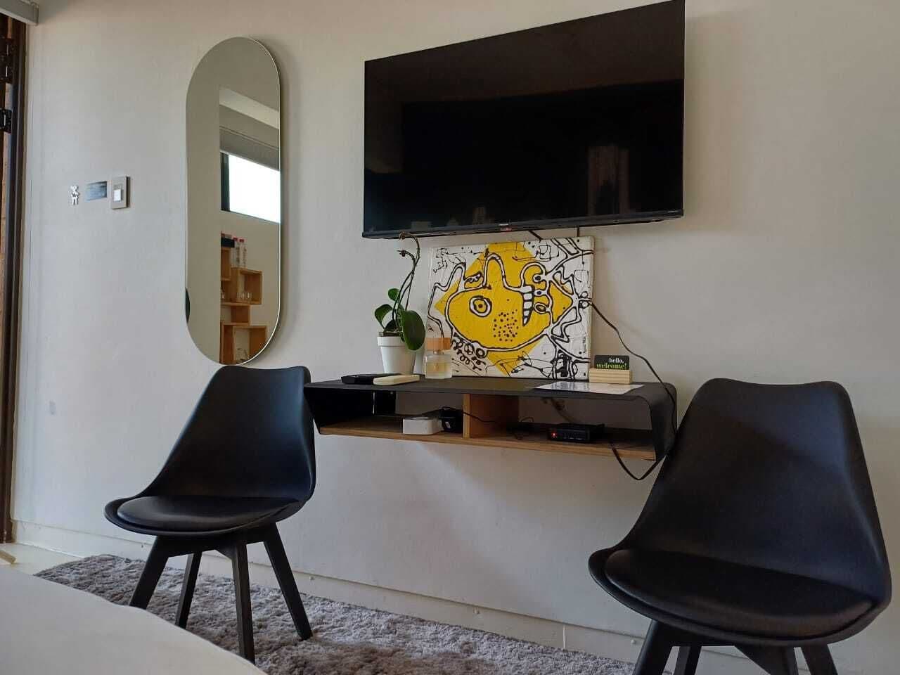 Una sala moderna con un televisor de pantalla plana montado en la pared, dos sillas negras, una mesita y un objeto decorativo con un personaje amarillo.