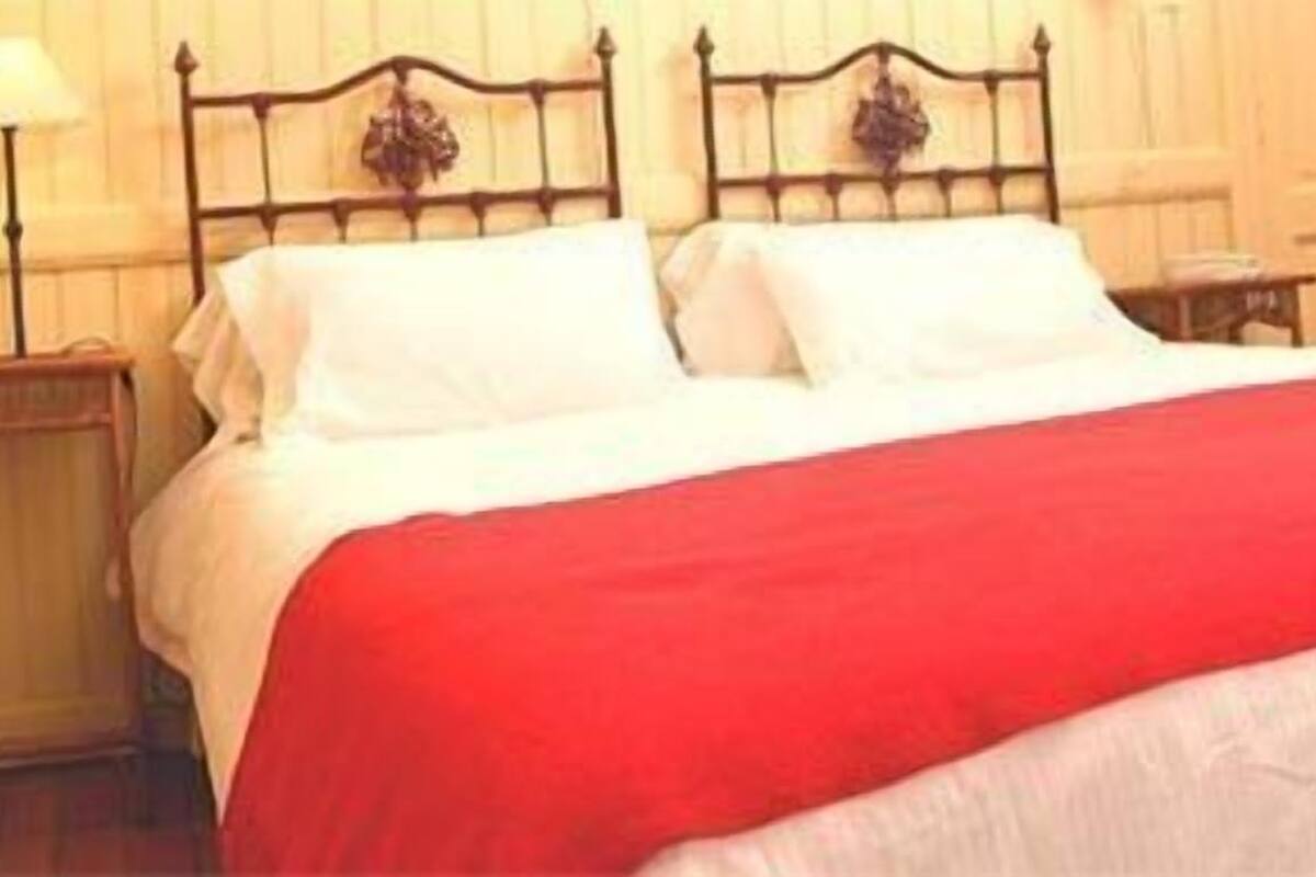 A bed with a red blanket and white pillows in a room with wooden walls and a nightstand with a lamp.
