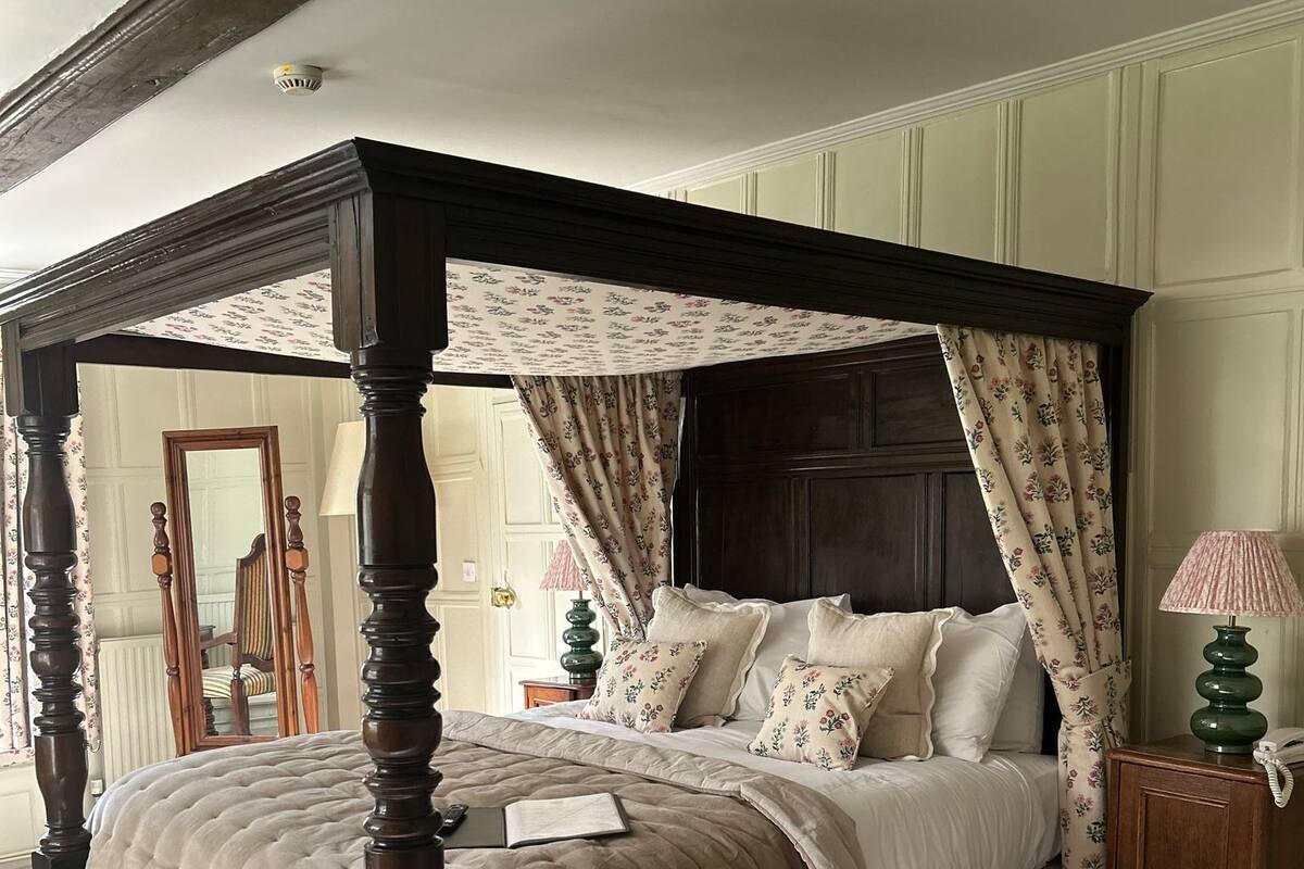 Un lit à baldaquin à quatre poteaux, surmonté d’un dais à motifs floraux, avec une lampe de chevet et une table de chevet dotée d’un téléphone.