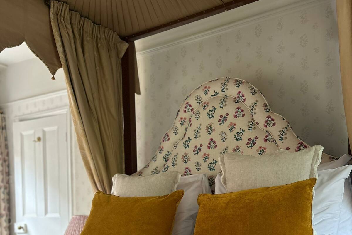 Un lit à baldaquin, avec une tête de lit ornée de motifs floraux et deux coussins de couleur moutarde.