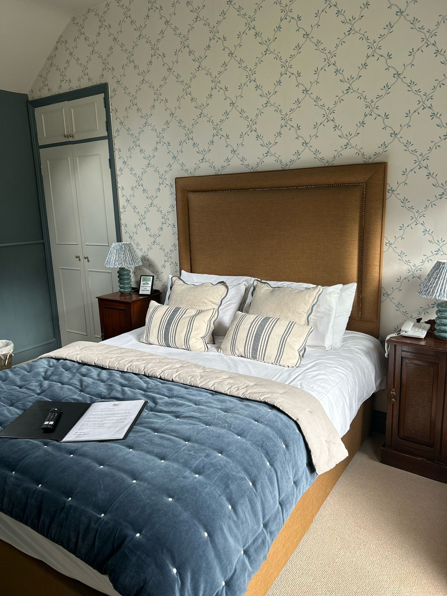 Een net opgemaakt bed met een blauwe quilt, een houten hoofdbord en gestreepte kussens. Er staat een nachtkastje met een lamp en een telefoon.
