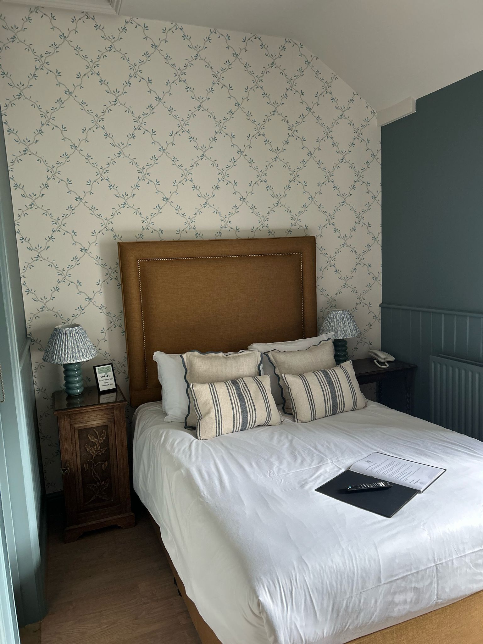 Una cama bien hecha con sábanas blancas y almohadas a rayas, cabecera de madera, mesita de noche con lámpara y un pequeño aparador decorativo.