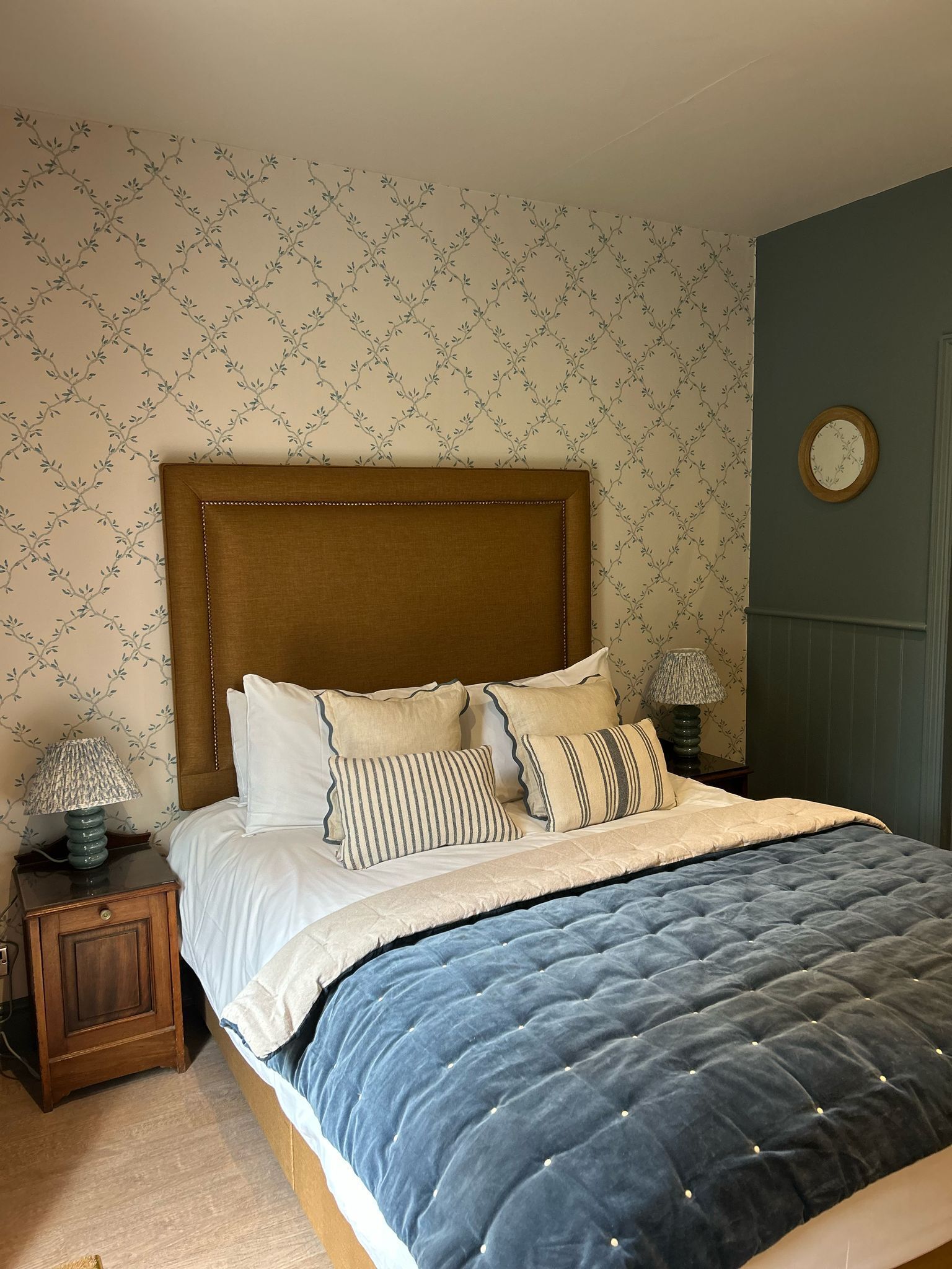 Un lit bien fait, avec une couette bleue, des oreillers blancs et une tête de lit en bois. On aperçoit une petite table de chevet avec une lampe, et une horloge est accrochée au mur.