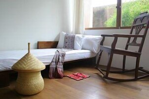 Um quarto pequeno e simples, com uma cama, uma cadeira de balanço e um banquinho de fibra.