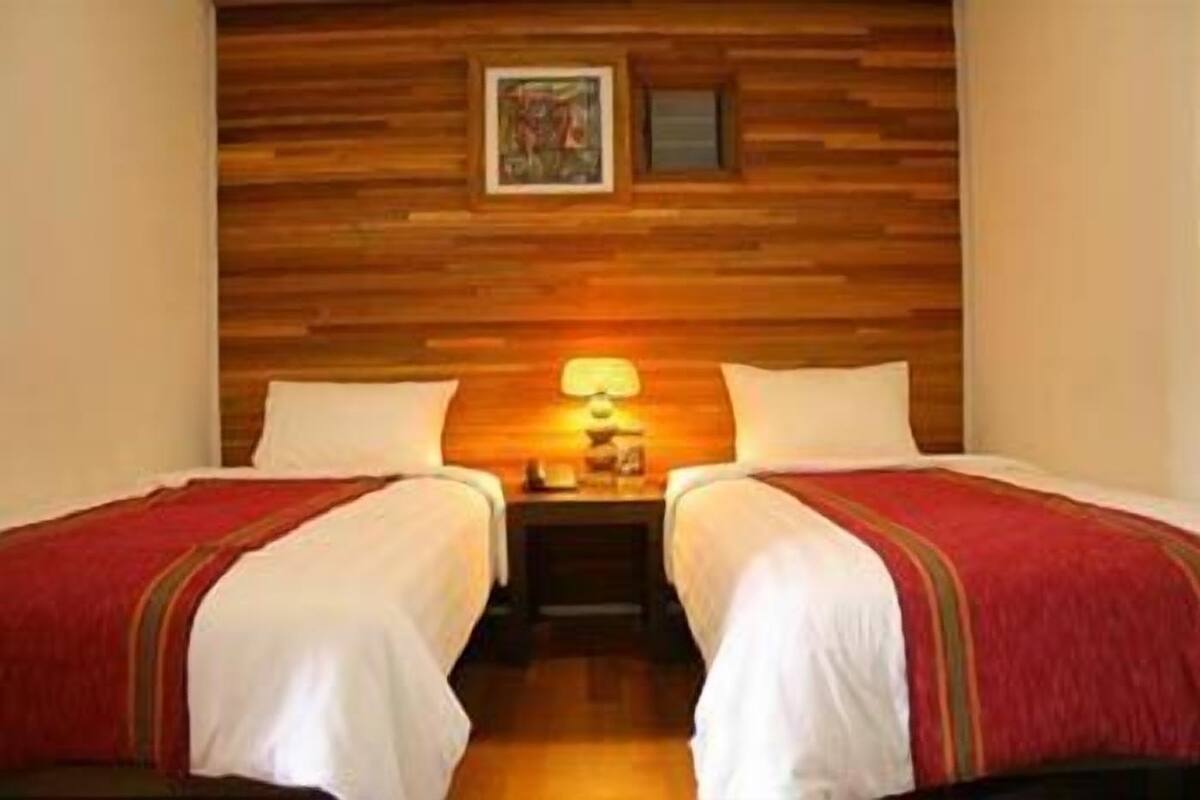 A room with two beds, wooden headboard, a nightstand with a lamp, and a framed picture on the wall.