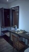 Um quarto pequeno e com pouca luz, com uma escrivaninha, uma cadeira e um espelho. Há uma mesinha com alguns objetos, incluindo uma garrafa e uma xícara.