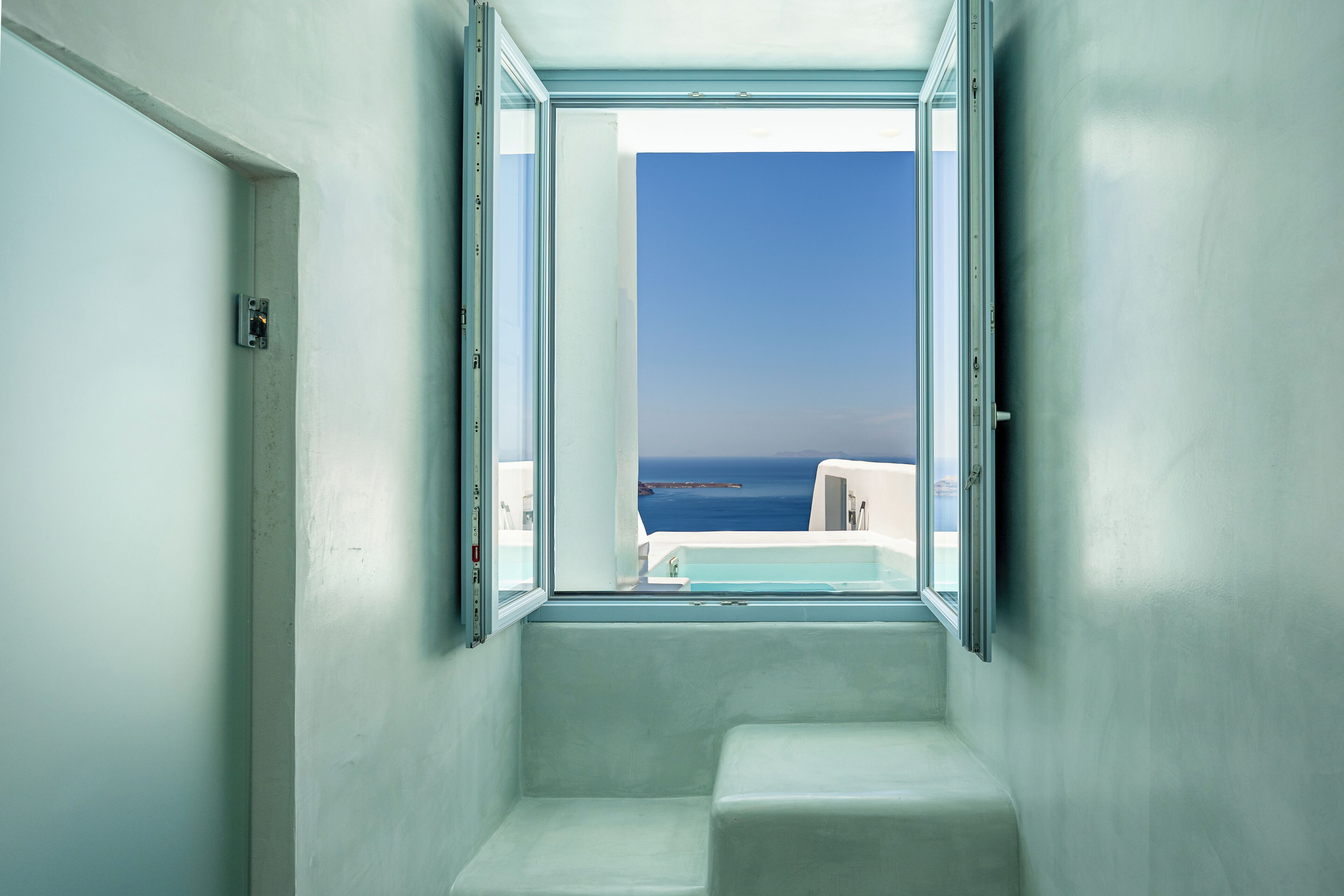 A narrow, white-walled bathroom with a small window overlooking a clear blue sky and a distant coastline.