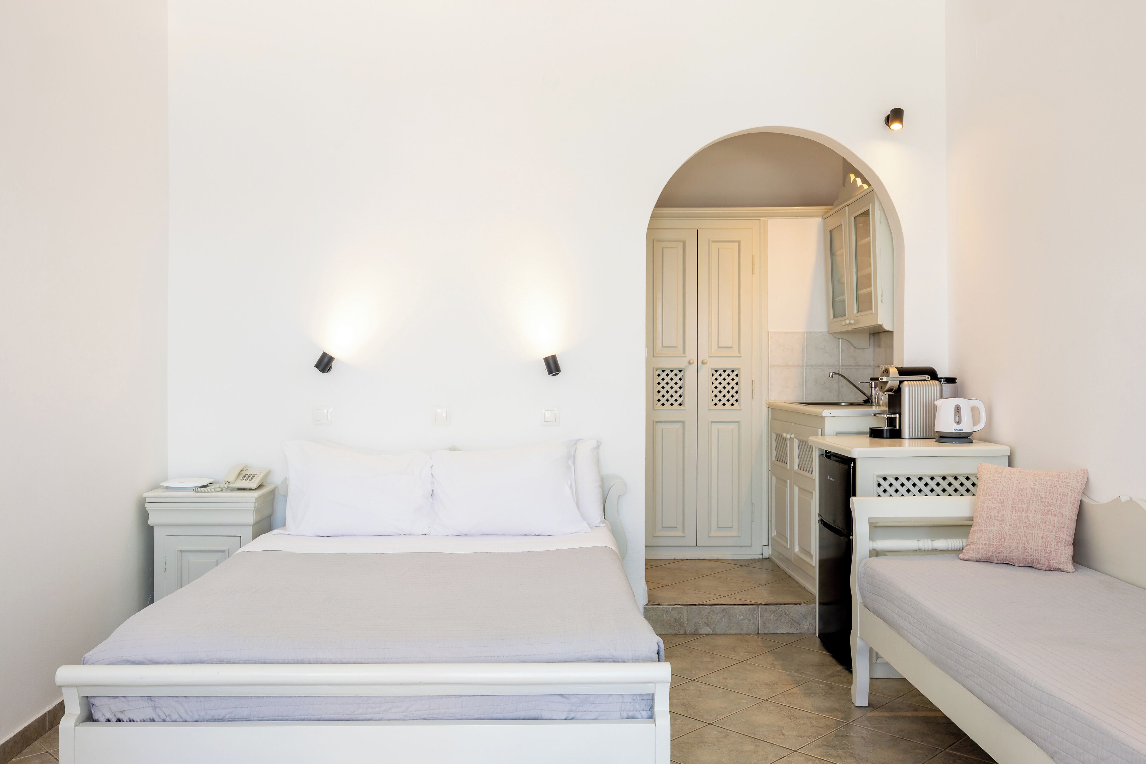 A bedroom with a bed, bedside tables, a nightstand, a dresser, a small kitchenette, and a bathroom.