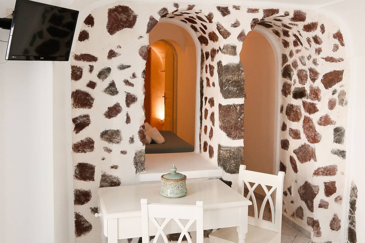 A room with a stone wall, a white table with chairs, a TV mounted on the wall, and a small window.