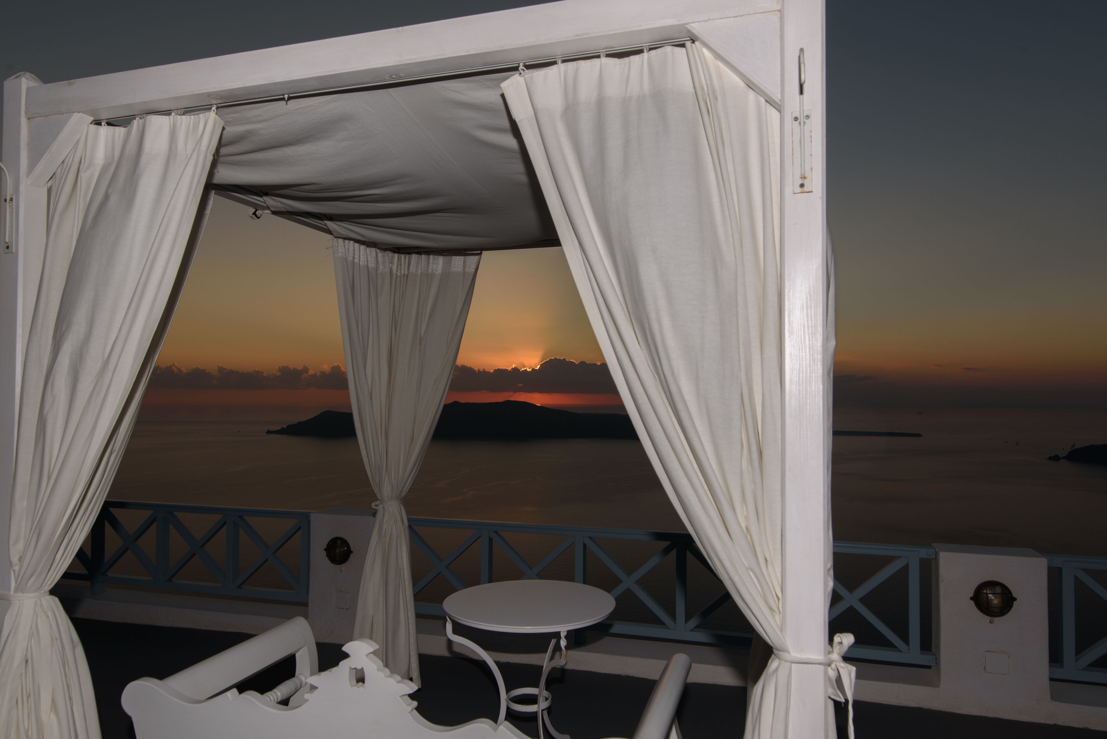 A balcony with a view of the sunset over the water, featuring a small round table and white curtains.