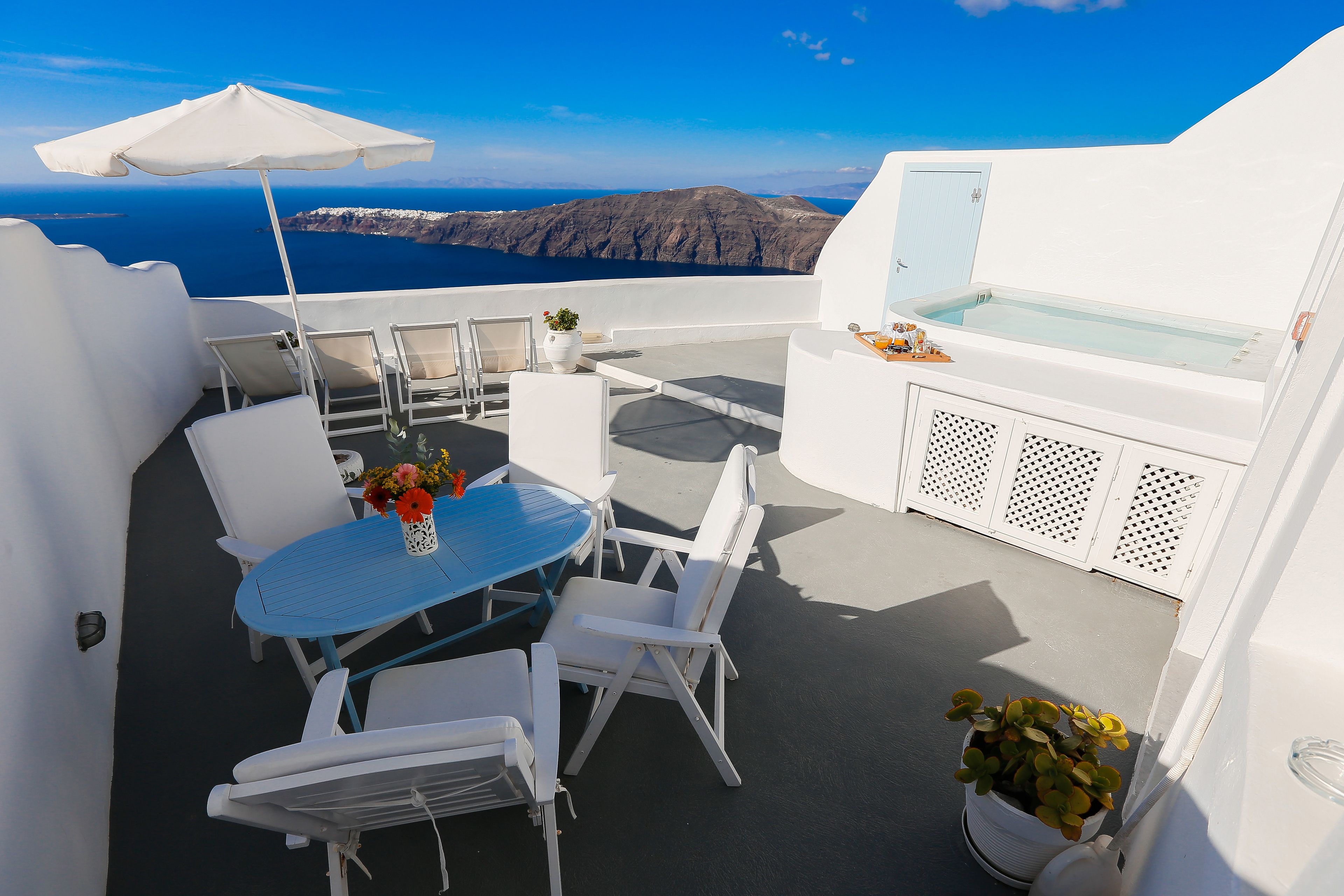 A white outdoor seating area with a table and chairs, a potted plant, and a view of the sea and mountains.