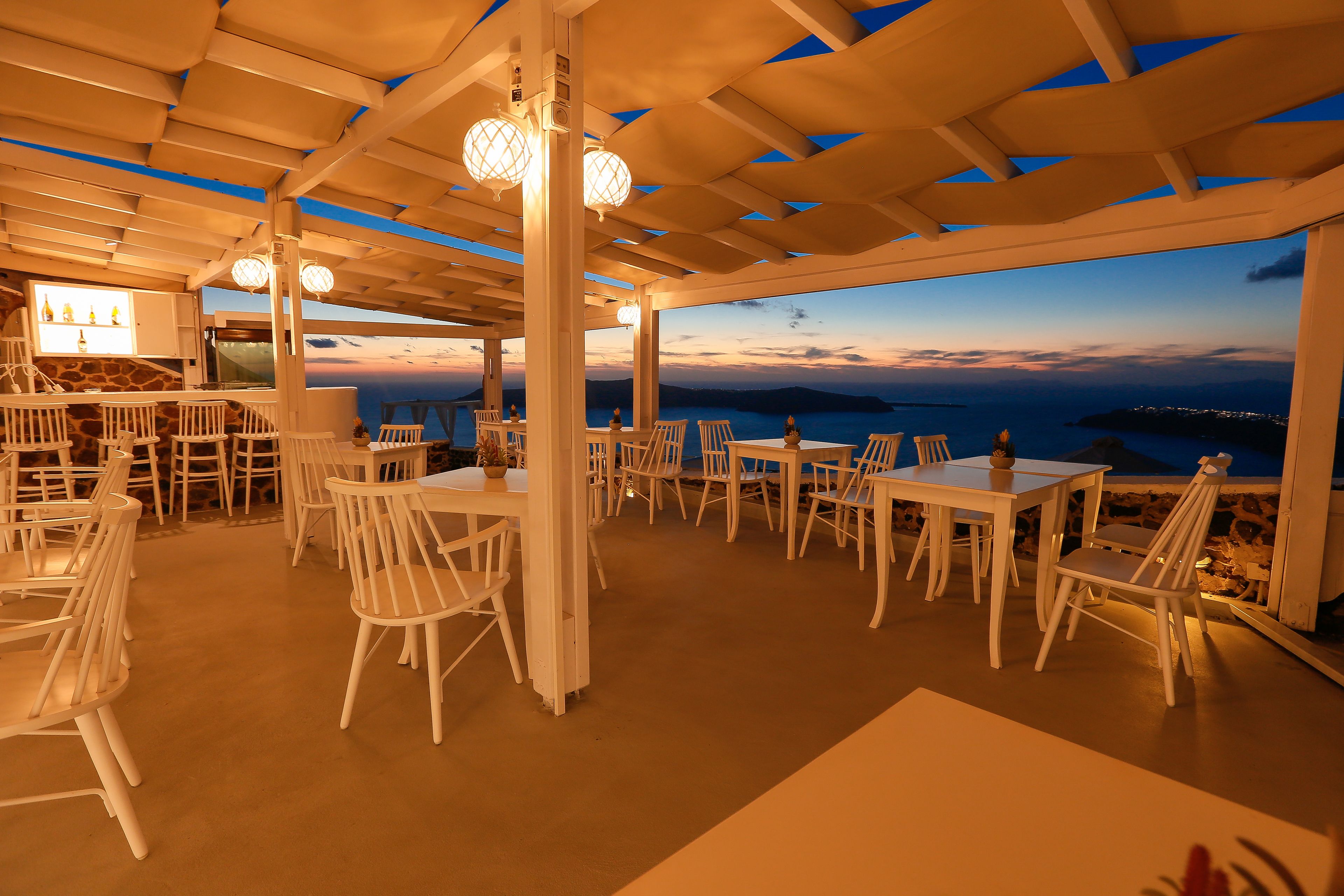 An outdoor dining area with white chairs and tables, a bar area, and a view of the sea at dusk.