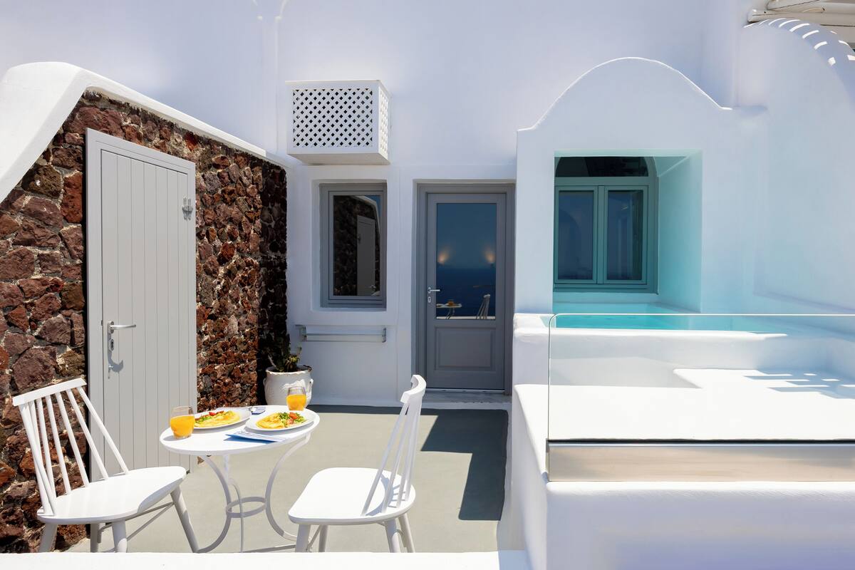 A white room with a stone wall, a door, and a window overlooking the sea.