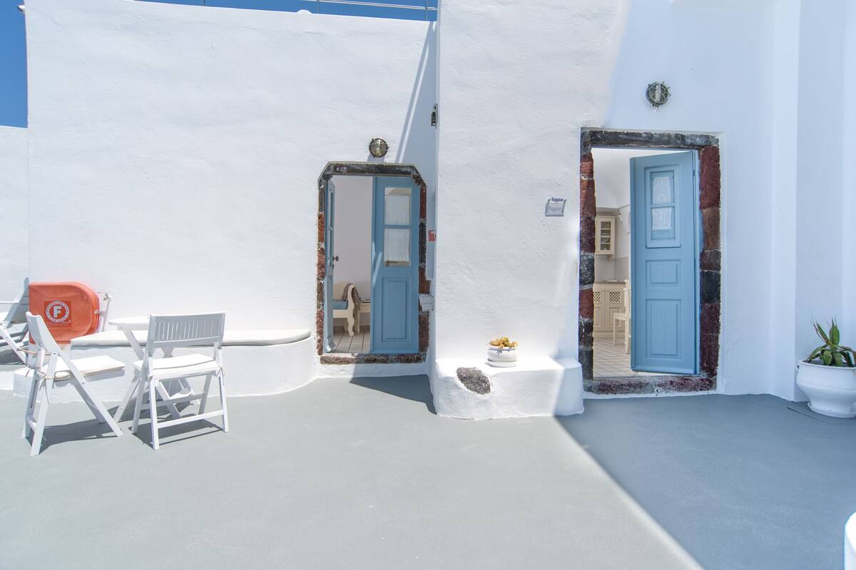 A white-walled building with two blue doors, a small table with a potted plant, and a white chair.