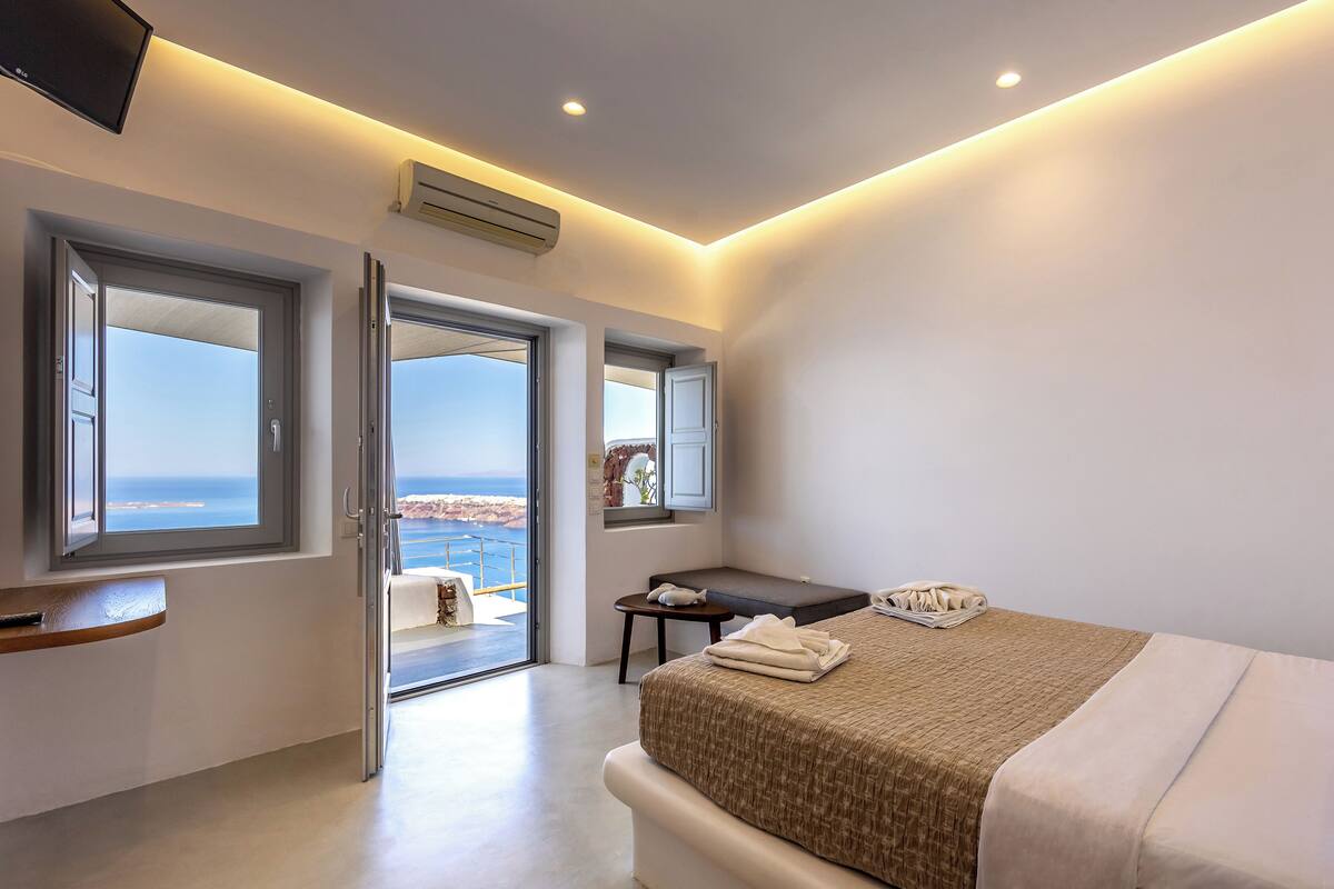 A modern bedroom with a large bed, a bedside table, and a view of the sea through open doors.