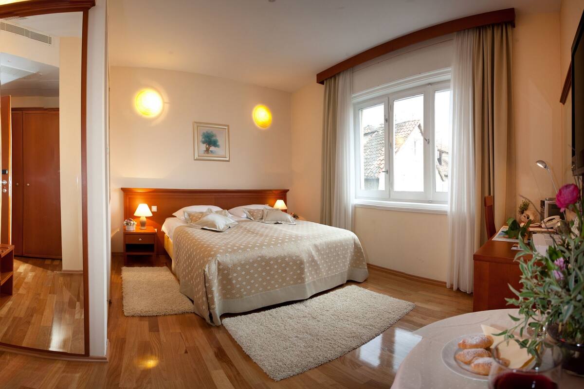 Une chambre à coucher avec une armoire en bois, un lit recouvert d’une couvre-lit à motifs, une table de chevet avec une lampe et une fenêtre avec des rideaux.