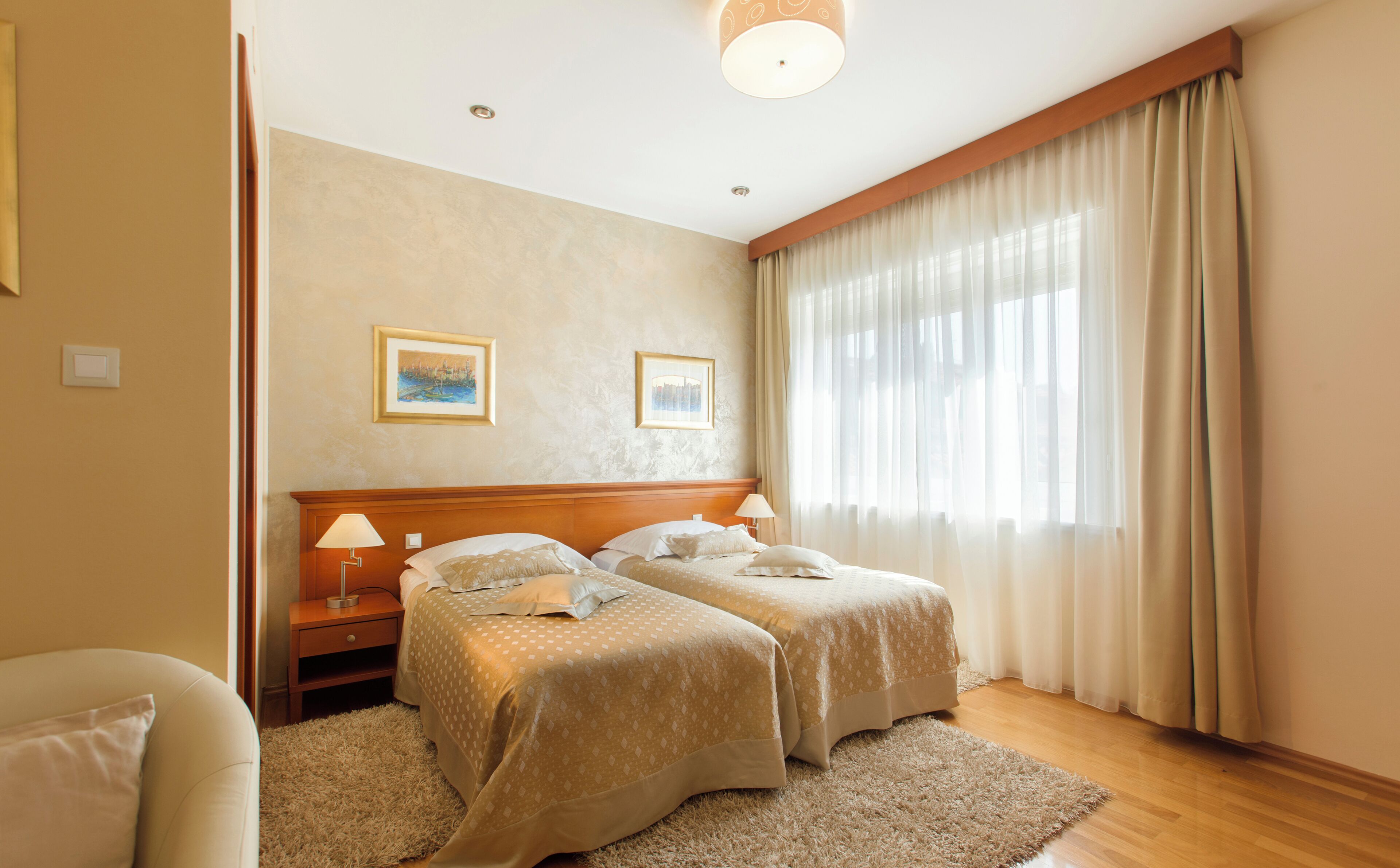 A hotel room with two beds, a wooden headboard, a large window with curtains, and two framed pictures on the wall.