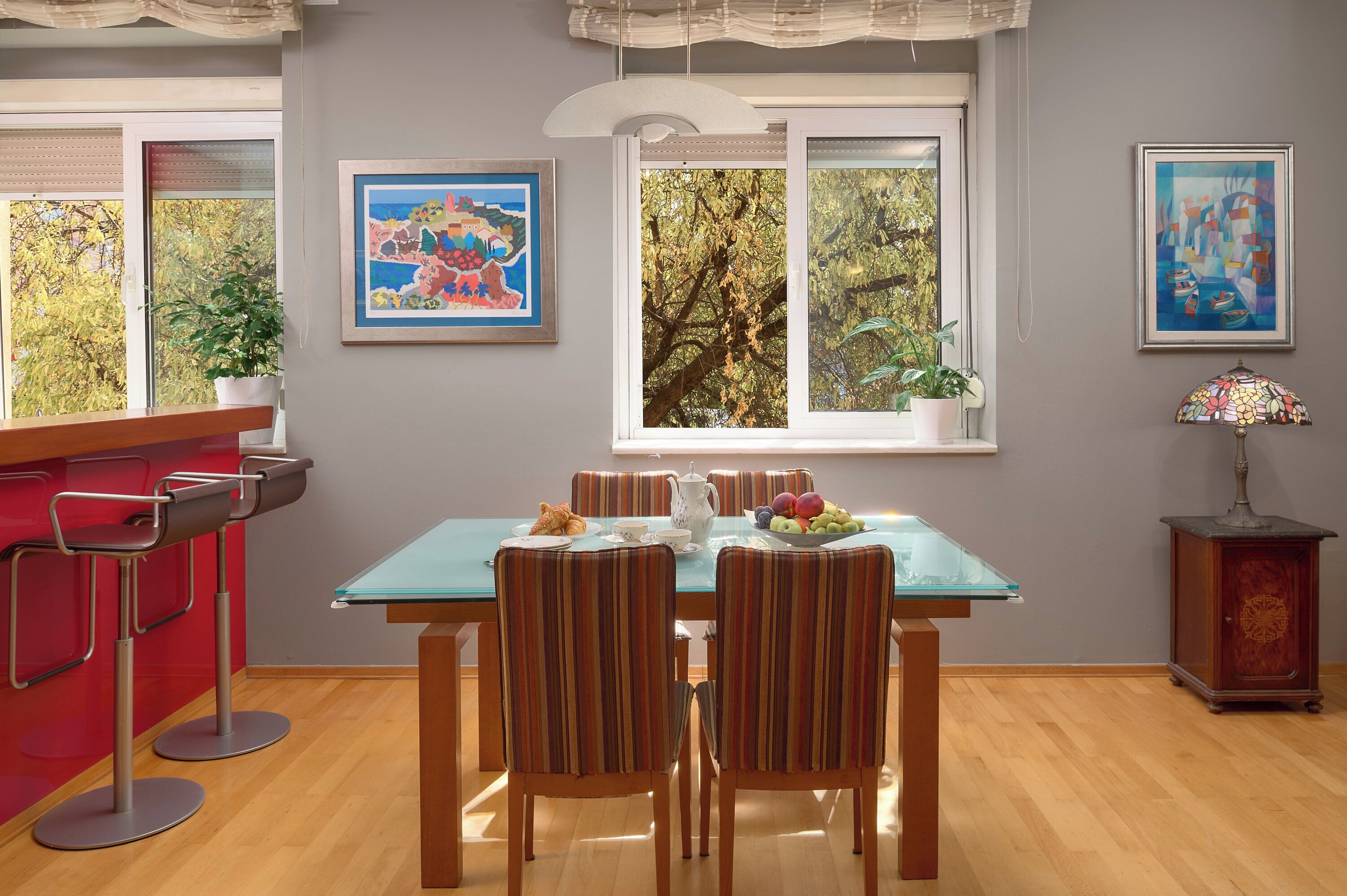 Un coin repas avec une table en verre, des chaises à rayures, un pichet blanc et un saladier rempli de fruits. Il y a deux fenêtres donnant sur des arbres et un tableau accroché au mur.