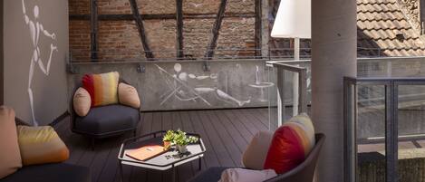 A rooftop terrace with a brick wall, a modern chair, a table with a plant, and a large lamp.