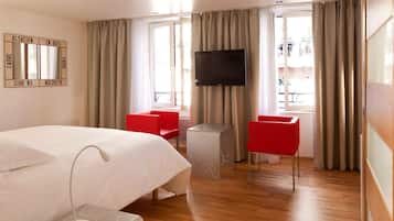 Habitació d'hotel amb un llit, dues cadires vermelles, una televisió i una finestra amb cortines.