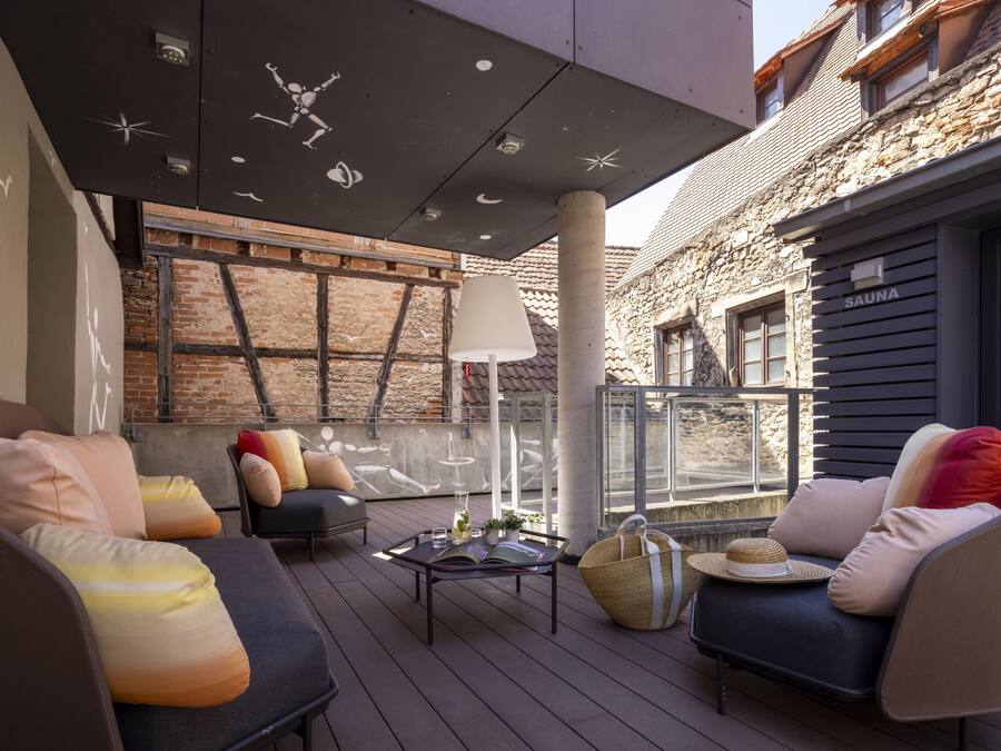 A modern outdoor seating area with cushioned chairs, a table with books and a drink, and a large window overlooking a stone building.