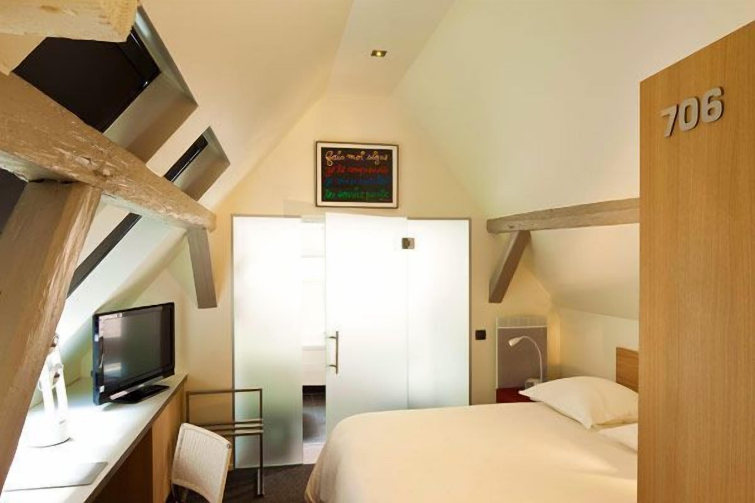 A hotel room with a bed, a desk with a television, a chair, a door, and a sign on the wall.