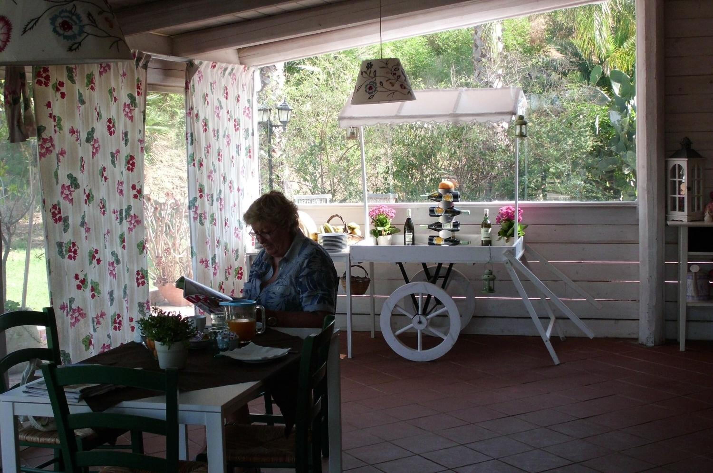 Un patio couvert avec une table dressée pour deux personnes, un chariot blanc rempli de bouteilles et une vue sur la verdure environnante.