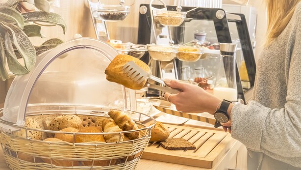 Montaje de bufé con una variedad de panes y pasteles en una cesta, una persona sirviendo comida y una vitrina con más opciones.