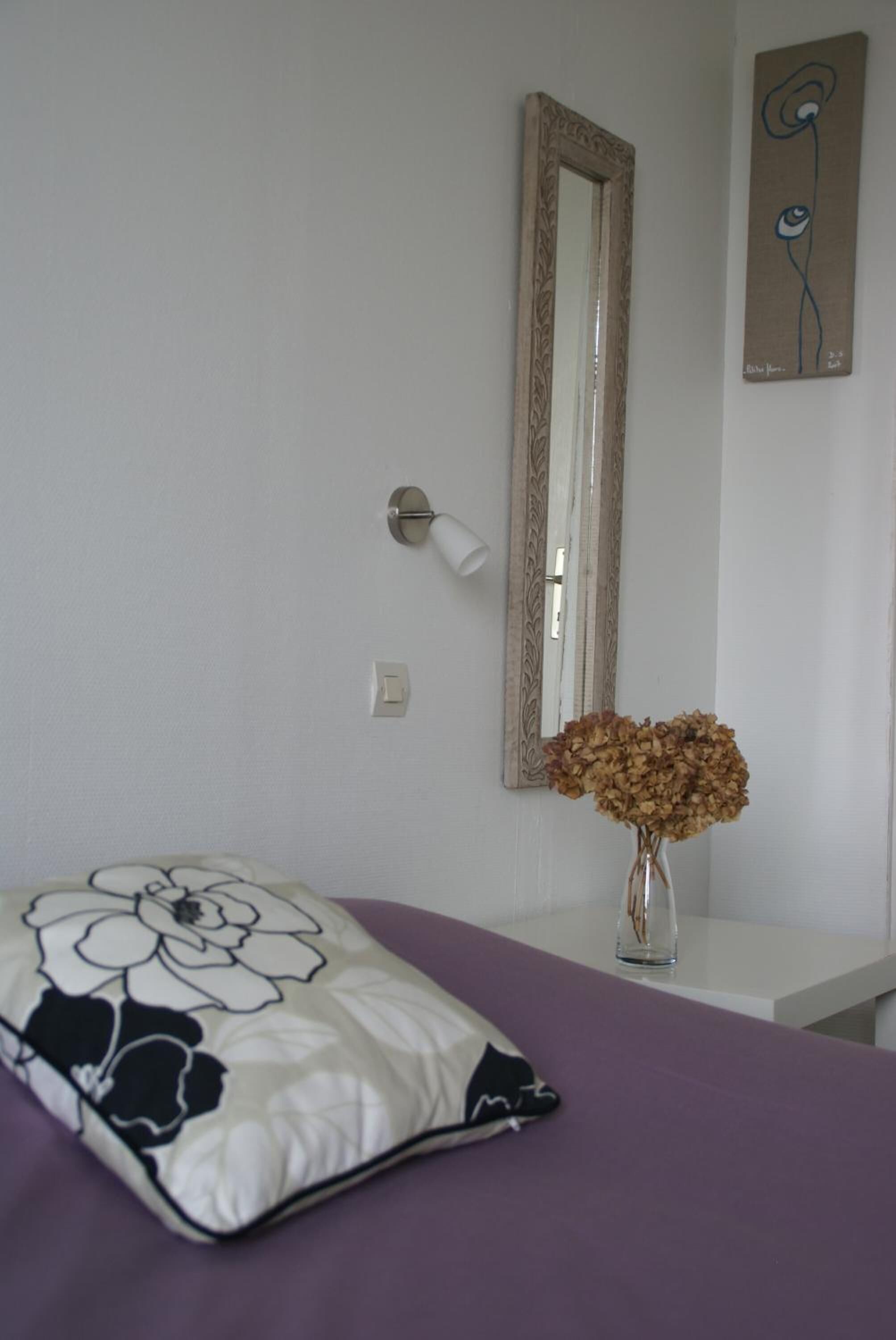 A bedroom with a bed, a bedside table with a vase of dried flowers, a wall-mounted mirror, and a painting.
