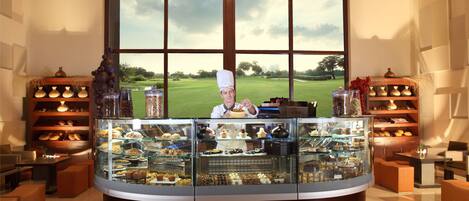 A bakery counter with a chef, various baked goods, and a view of a golf course through large windows.