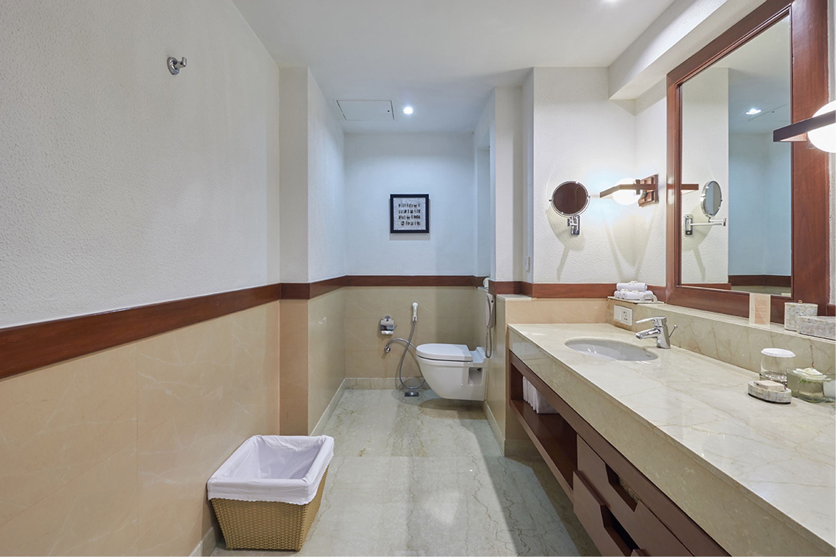 A bathroom with a toilet, bidet, sink, and mirror. There is a trash bin and a framed sign on the wall.