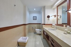 A bathroom with a toilet, bidet, sink, and mirror. There is a trash bin and a framed sign on the wall.
