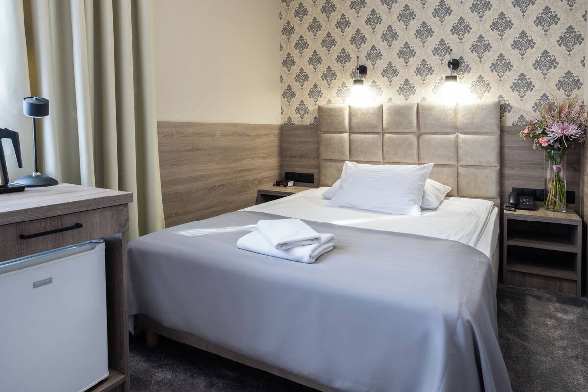 A hotel room with a bed, bedside tables, a lamp, a nightstand, and a small refrigerator.