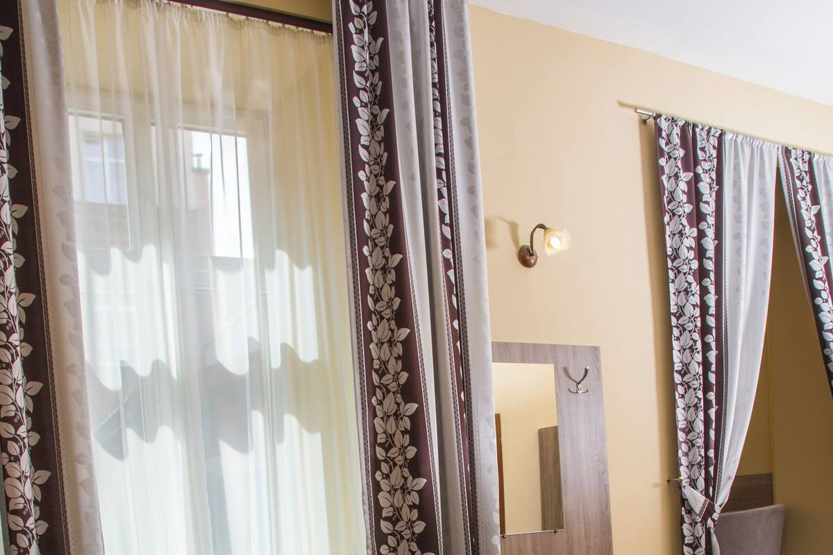 A hotel room with a bed, a window with sheer curtains, a bedside table with a kettle and bottles, and a mirror on the wall.