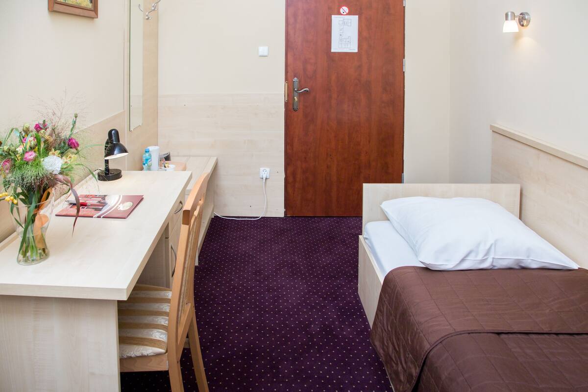 A small, neatly arranged room with a wooden desk, a chair, a bed with a brown bedspread, and a purple carpet.