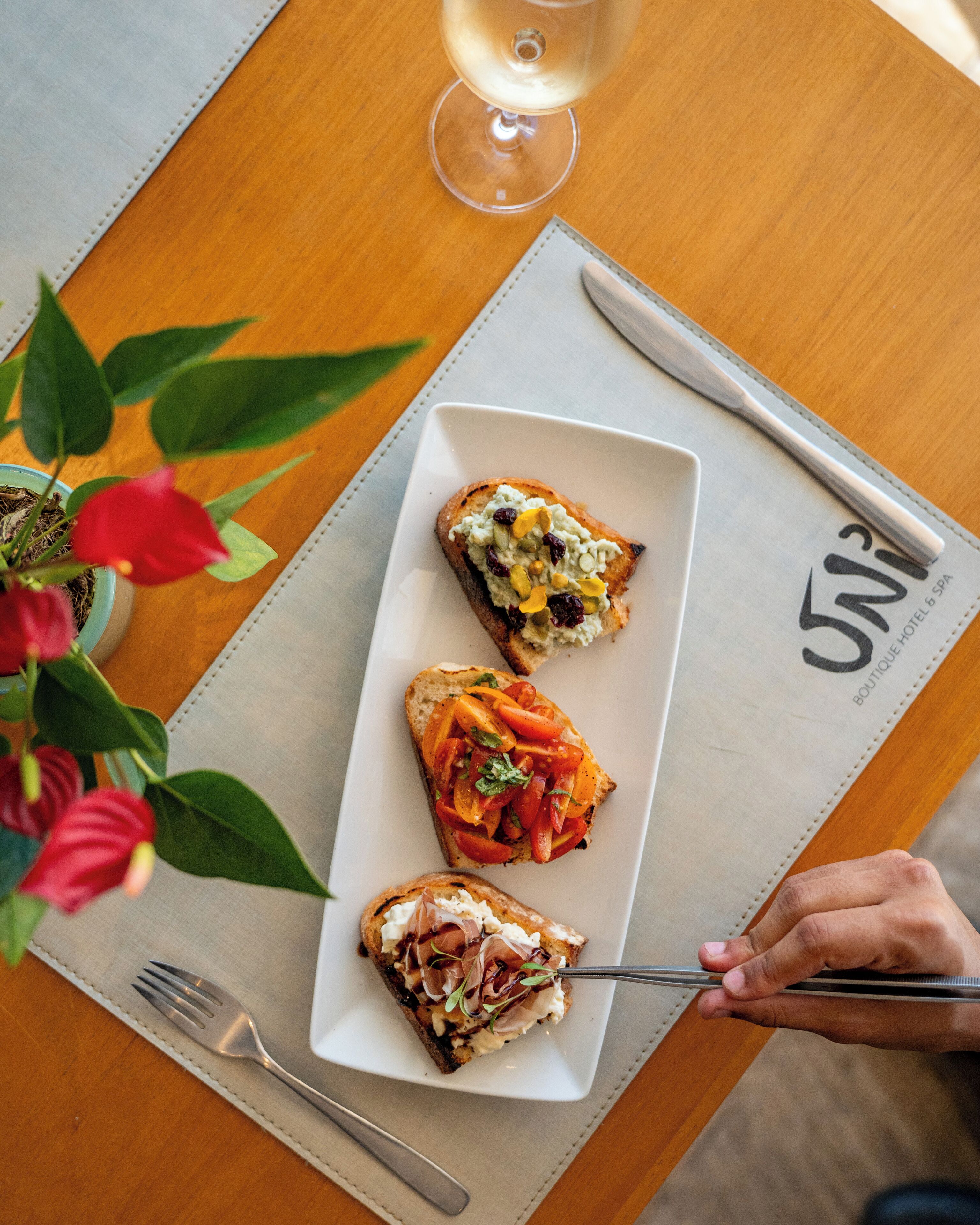 Una mesa con un plato de comida, una copa de vino blanco y una mano sosteniendo un tenedor.